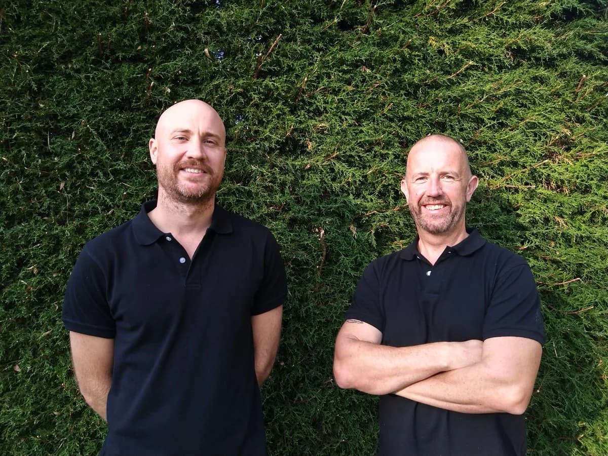 ABS Horticulture team members Anthony and Sam standing together in matching black polo shirts in front of a green hedge